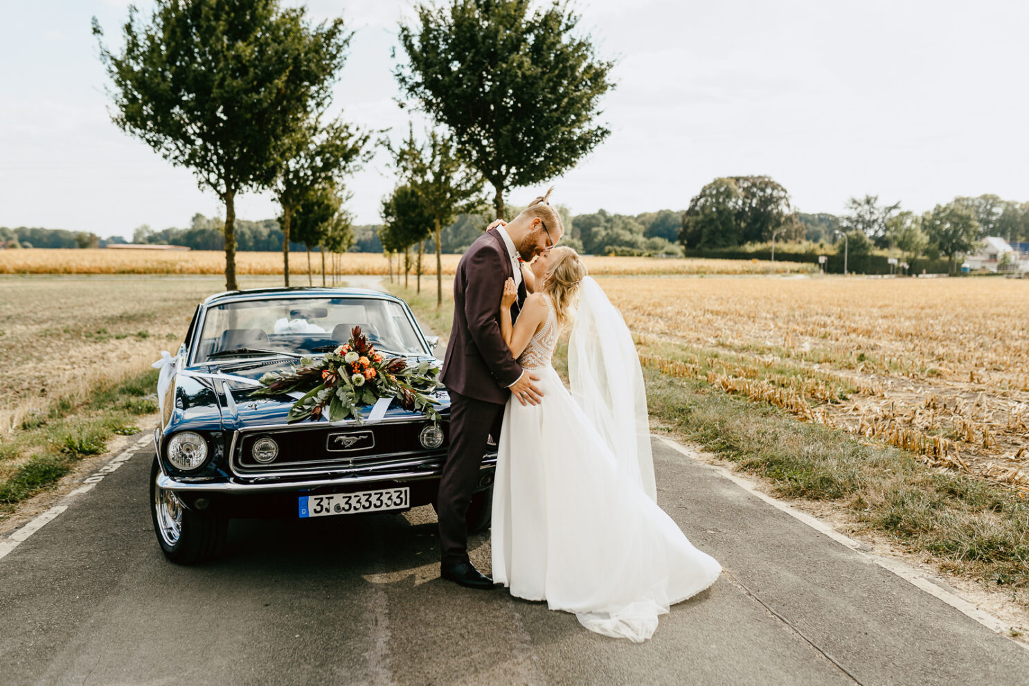 Hochzeitspaar küsst sich auf Allee vor blauem Ford Mustang von Hochzeitsfotograf Bielefeld