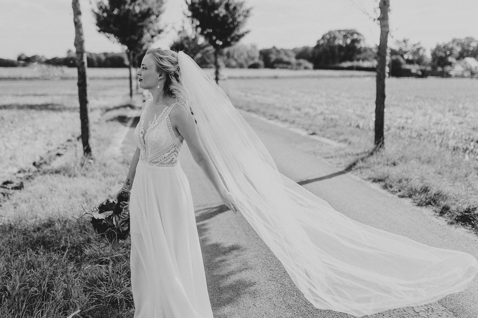 Schwarz Weiß Foto von Braut auf Allee, der Schleier weht im Wind, nach Hochzeit im Bockhorster Kotten in Verl