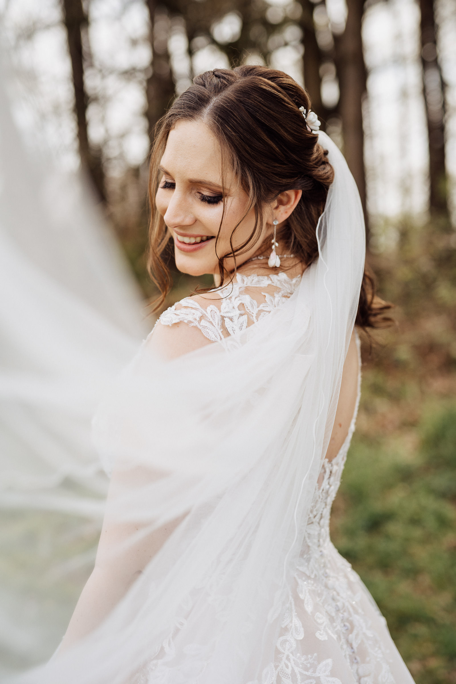 Es ist eine Braut von hinten zu sehen. Sie steht im Wald und lächelt über ihre linke Schulter Richtung Kamera. Im Vordergrund ist ihr Schleier zu sehen, der im Wind fliegt.