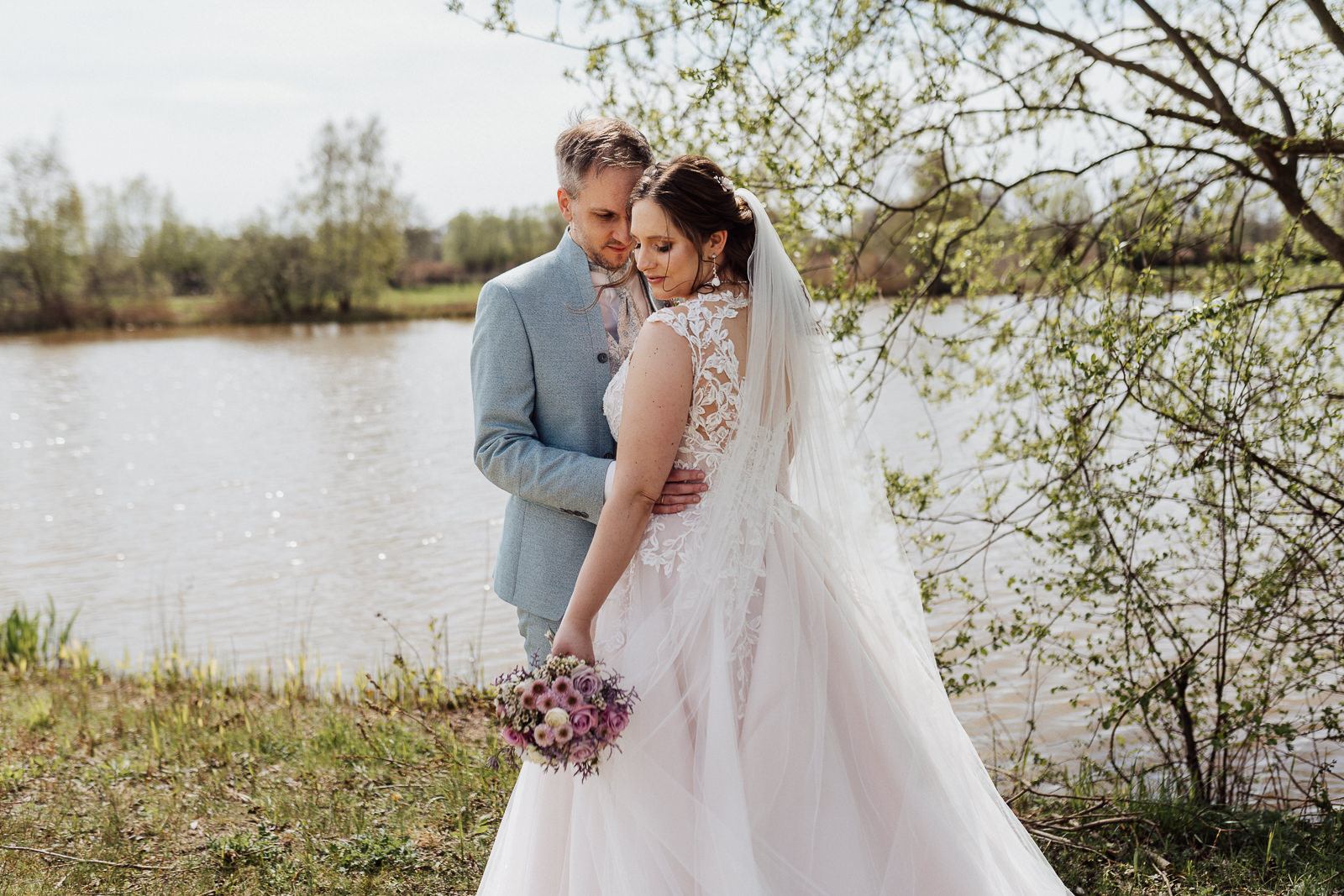 Hochzeitsfoto von einem Brautpaar vor einem See, beiden stehen voreinander und lehnen die Köpfe aneinander.
