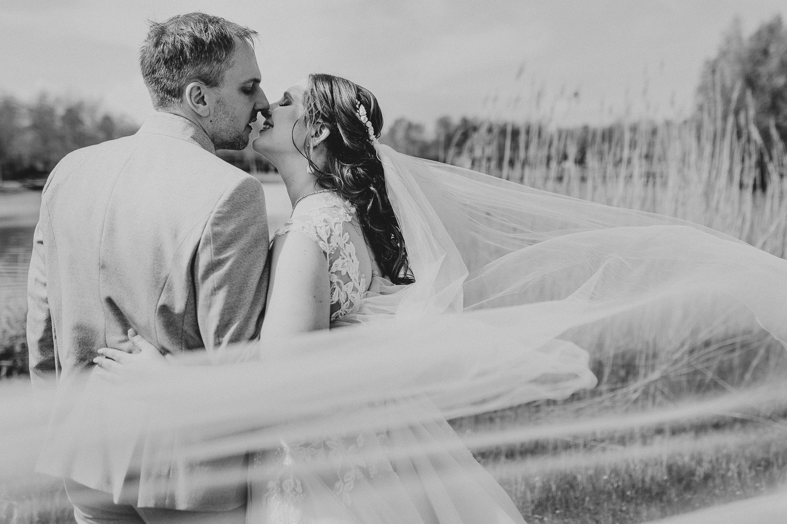 Ein schwarz weiß Foto von einem Brautpaar. Es ist von hinten zu sehen und steht an einem See. Die beiden küssen sich, der Schleier fliegt im Wind im Vordergrund bei einer Hochzeit im Lind am See in Rietberg.