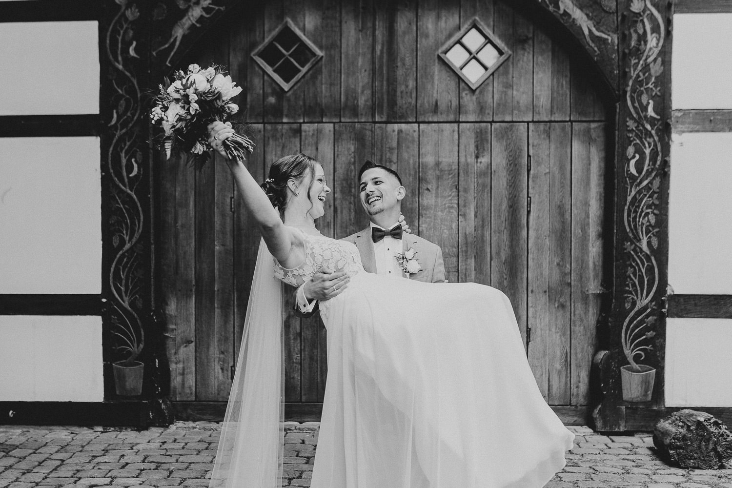 Hochzeitsfoto vor Fachwerkhaus. Der Bräutigam trägt seine Braut auf den Armen, die Braut hält ihren Brautstrauß in die Luft.